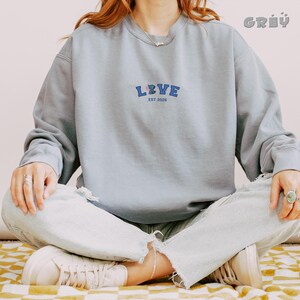 May include: A light grey sweatshirt with the word "LOVE" embroidered in blue and pink, with "EST 2026" underneath. The sweatshirt is worn with light-wash jeans and white sneakers. The word "GREY" is printed in the top right corner.