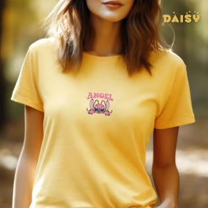 Pode incluir: Camiseta amarela mostarda com um desenho de personagem de desenho animado. O desenho apresenta um personagem rosa e roxo com a palavra "ANGEL" acima. A camiseta tem mangas curtas e gola redonda. O desenho &eacute; centrado no peito.