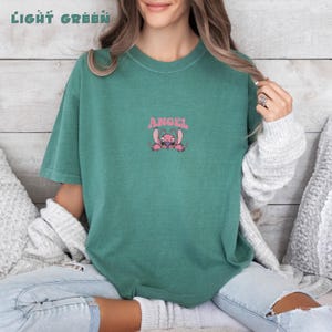 Puede incluir: Camiseta verde claro con la palabra "ANGEL" sobre un personaje de dibujos animados rosa. La camiseta es de color liso y tiene mangas cortas. Las palabras "LIGHT GREEN" est&aacute;n en la parte superior de la imagen.
