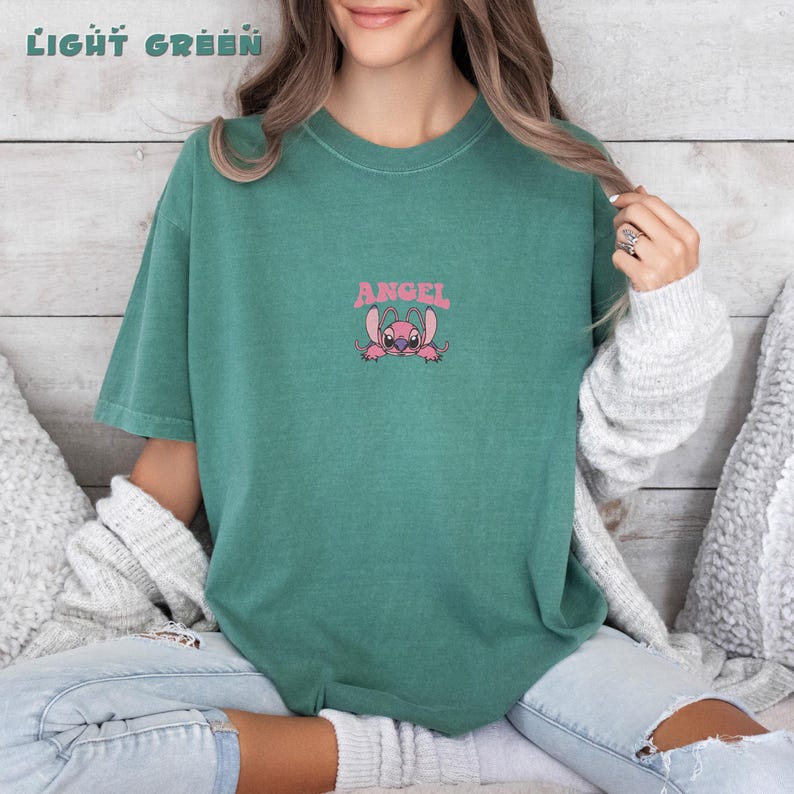 Puede incluir: Camiseta verde claro con la palabra "ANGEL" sobre un personaje de dibujos animados rosa. La camiseta es de color liso y tiene mangas cortas. Las palabras "LIGHT GREEN" est&aacute;n en la parte superior de la imagen.