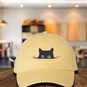 May include: A light yellow baseball cap featuring a black cat peeking over the brim. The cat's eyes and whiskers are visible. The cap has a curved brim and a button on top.