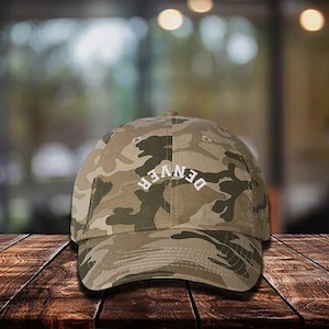 May include: Camouflage baseball cap with the word "DENVER" embroidered in white across the front. The cap features a curved brim and a classic six-panel design. The hat is made of a durable fabric.