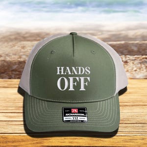 Pu&ograve; includere: Cappellino da camionista verde oliva e bianco con la scritta "HANDS OFF" ricamata in bianco. Il cappellino ha una visiera curva e un cinturino regolabile. L'etichetta del marchio Richardson &egrave; visibile.