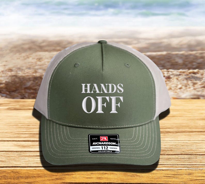 Pu&ograve; includere: Cappellino da camionista verde oliva e bianco con la scritta "HANDS OFF" ricamata in bianco. Il cappellino ha una visiera curva e un cinturino regolabile. L'etichetta del marchio Richardson &egrave; visibile.