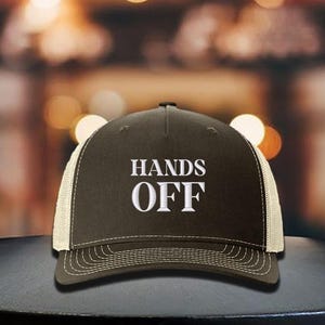 Pu&ograve; includere: Cappellino da camionista marrone con retro in rete bianca e visiera curva. La parte anteriore del cappellino presenta la scritta ricamata "HANDS OFF" in lettere bianche. Il cappellino &egrave; su una superficie scura.
