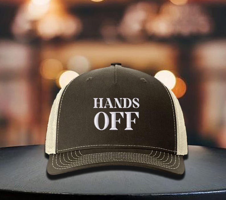 Pu&ograve; includere: Cappellino da camionista marrone con retro in rete bianca e visiera curva. La parte anteriore del cappellino presenta la scritta ricamata "HANDS OFF" in lettere bianche. Il cappellino &egrave; su una superficie scura.