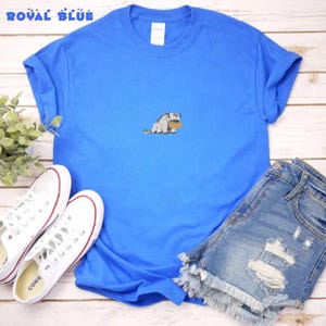May include: A royal blue short-sleeve t-shirt featuring a small, centered embroidered graphic of a light gray, furry creature with brown horns and a brown saddle. The shirt has a crew neckline and is made of a solid, smooth fabric.