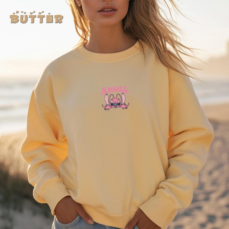 Pode incluir: Um moletom amarelo claro com a palavra "BUTTER" em letras douradas no canto superior esquerdo. O moletom apresenta um personagem de desenho animado rosa com a palavra "ANGEL" acima. A pessoa est&aacute; em uma praia.