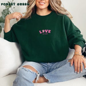 May include: A forest green crew neck sweatshirt featuring the word "LOVE" in pink with a floral design and "EST 2026". The text "FOREST GREEN" is at the top. The sweatshirt is a casual style.