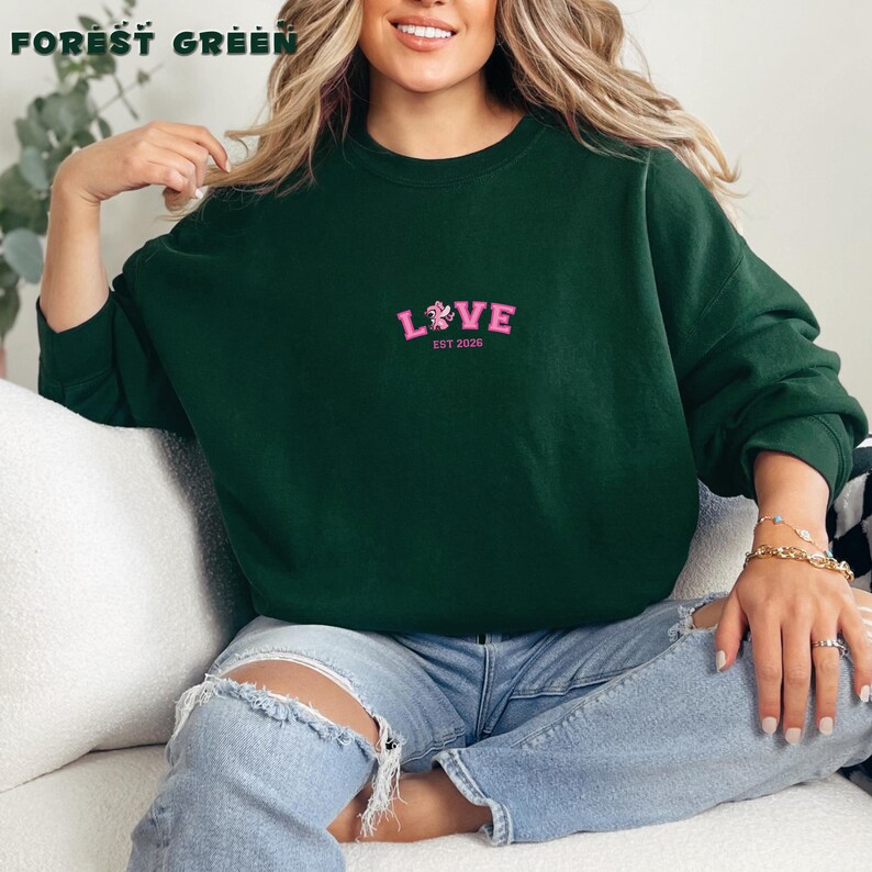 May include: A forest green crew neck sweatshirt featuring the word "LOVE" in pink with a floral design and "EST 2026". The text "FOREST GREEN" is at the top. The sweatshirt is a casual style.