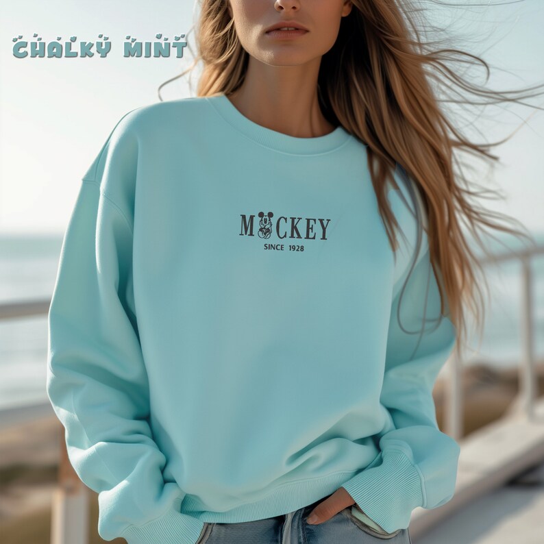 May include: A light blue sweatshirt with the word "Mickey" and the phrase "Since 1928" printed in black. The sweatshirt is a solid color and has a crew neck. The brand name "Chalky Mint" is printed at the top.