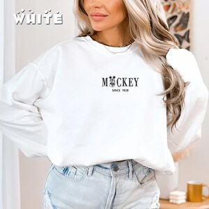 May include: White sweatshirt with the word "WHITE" in a stylized font at the top left. The sweatshirt features a black graphic of Mickey Mouse with the word "MOCKEY" and "SINCE 1928" printed below.