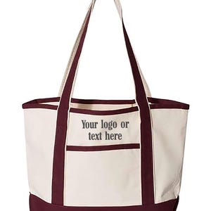 May include: Cream-coloured tote bag with burgundy accents. Features a burgundy base, trim, and handles. A front pocket is outlined in burgundy. The text "Your logo or text here" is printed on the front of the bag.