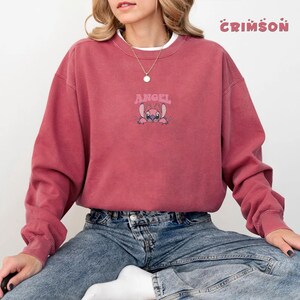Pode incluir: Um moletom carmesim com a palavra "ANGEL" acima de um personagem de desenho animado rosa. O moletom tem mangas compridas e gola redonda. A palavra "CRIMSON" &eacute; impressa no canto superior direito.