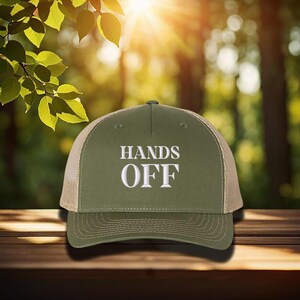 Pu&ograve; includere: Cappellino da camionista verde oliva con la scritta bianca "HANDS OFF". Il cappellino ha il retro in rete e una visiera curva. Il cappellino &egrave; appoggiato su una superficie di legno.