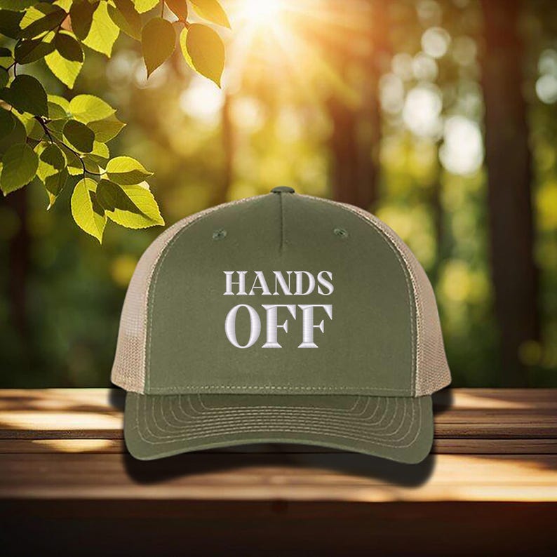 Pu&ograve; includere: Cappellino da camionista verde oliva con la scritta bianca "HANDS OFF". Il cappellino ha il retro in rete e una visiera curva. Il cappellino &egrave; appoggiato su una superficie di legno.