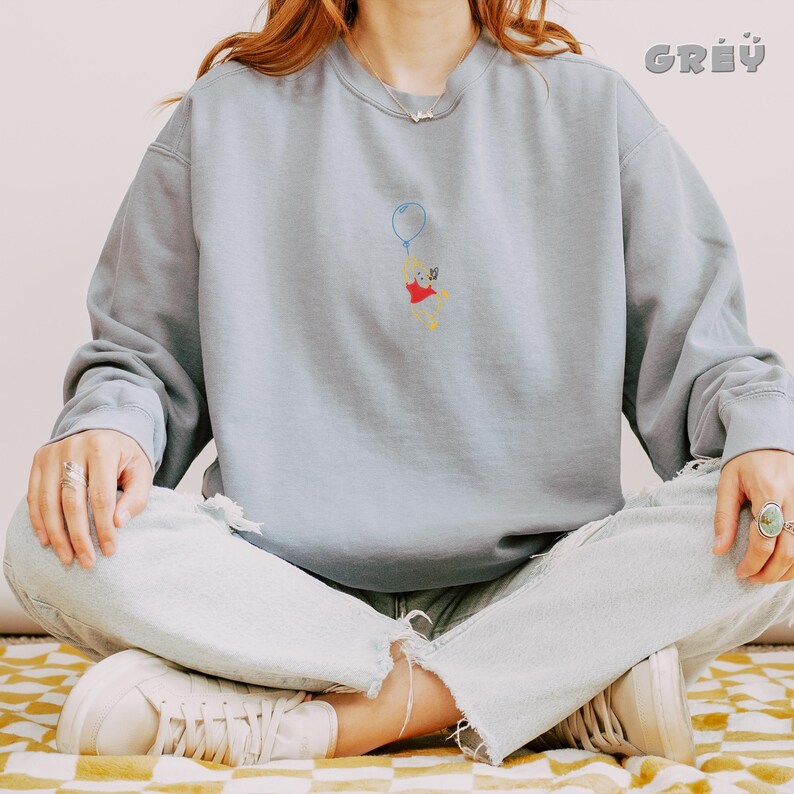 May include: A light blue sweatshirt with an embroidered image of a cartoon bear holding a balloon. The bear is wearing a red shirt and has a yellow face.