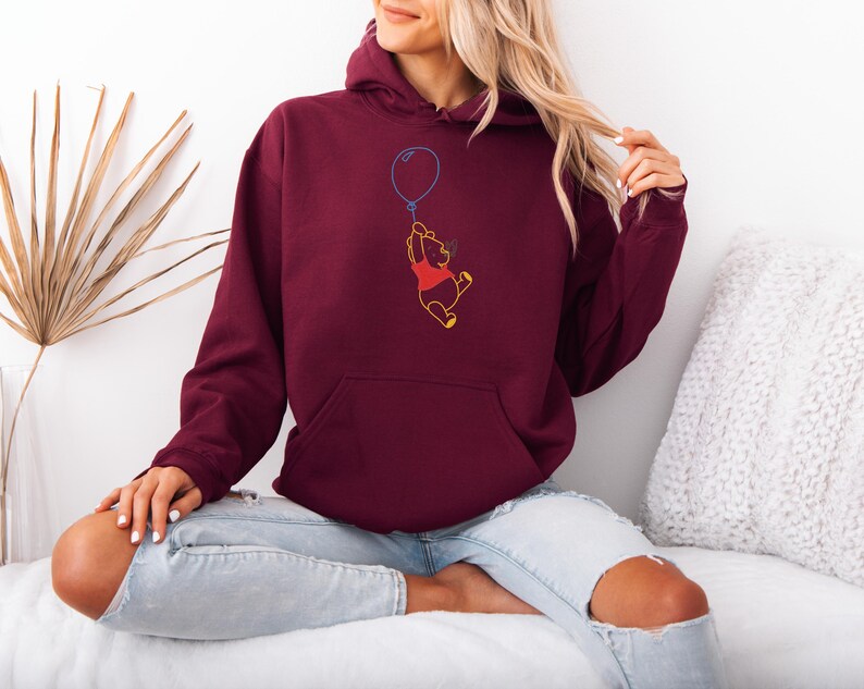 Puede incluir: Sudadera con capucha burdeos con bolsillo delantero y capucha con cord&oacute;n. La sudadera presenta un dise&ntilde;o bordado de un personaje amarillo de Winnie the Pooh sosteniendo un globo azul. El personaje lleva una camisa roja.