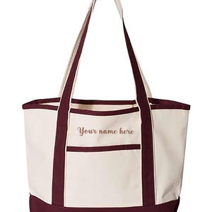 May include: Cream-colored tote bag with burgundy accents. Features a burgundy base, trim, and handles. A front pocket and the text "Your name here" are visible. The bag is designed for carrying items.