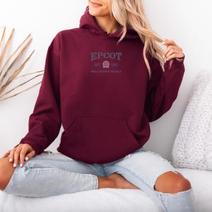 May include: A maroon hoodie with a front pocket and a hood. The text "EPCOT EST 1982 WALT DISNEY WORLD" is printed in navy blue. The person is wearing light blue jeans.