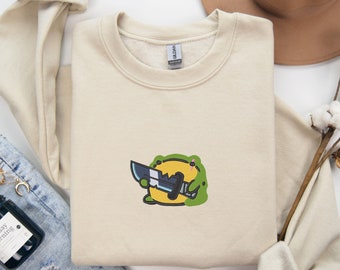May include: A beige sweatshirt with a green and yellow cartoon frog holding a knife. The frog is wearing a black eye patch. The sweatshirt has the word "SAND" printed on the top right corner.