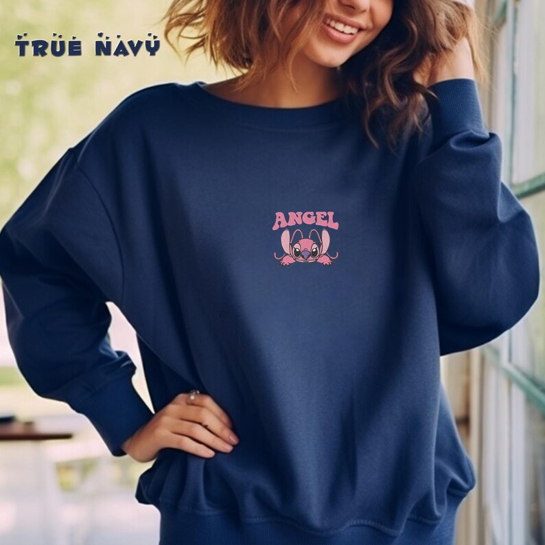 Pode incluir: Um moletom azul marinho com a palavra "ANGEL" acima de um personagem de desenho animado rosa. O moletom tamb&eacute;m tem as palavras "TRUE NAVY" impressas na parte superior. A parte superior de manga comprida &eacute; de cor s&oacute;lida.
