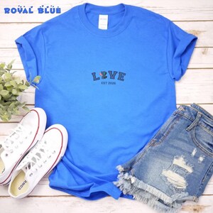 Puede incluir: Camiseta azul real con la palabra "LOVE" bordada en la parte delantera, junto con el texto "EST 2026". La camiseta se combina con pantalones cortos de mezclilla y zapatillas blancas. Las zapatillas tienen cordones blancos y detalles en rojo.