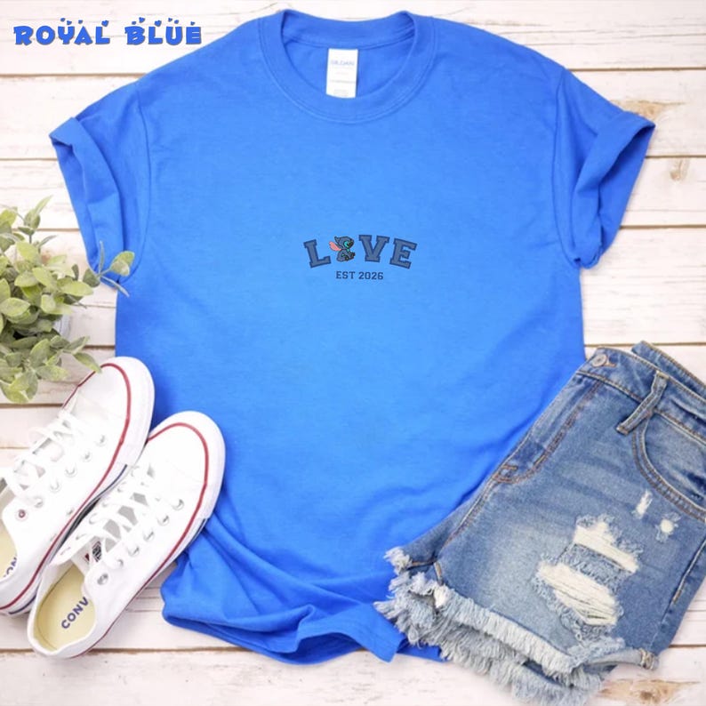 Puede incluir: Camiseta azul real con la palabra "LOVE" bordada en la parte delantera, junto con el texto "EST 2026". La camiseta se combina con pantalones cortos de mezclilla y zapatillas blancas. Las zapatillas tienen cordones blancos y detalles en rojo.
