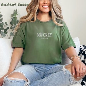 May include: A woman wearing a green t-shirt with the text "MICKEY SINCE 1928" and a small Mickey Mouse graphic. She is sitting on a white couch with her legs crossed.