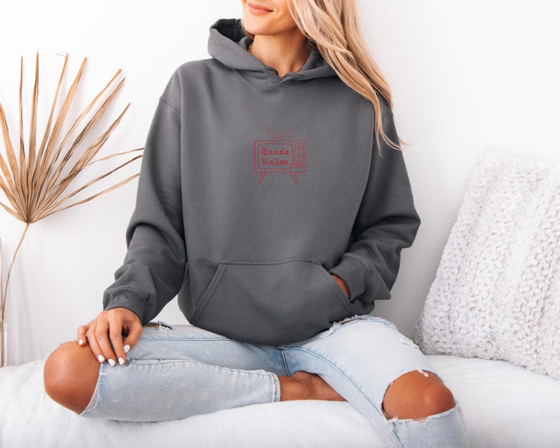 Pu&ograve; includere: Una felpa con cappuccio grigio scuro con una tasca frontale e un cappuccio. La felpa presenta una grafica rossa di un televisore retr&ograve; con le parole "Wanda Vision" all'interno. La modella indossa jeans blu chiaro.