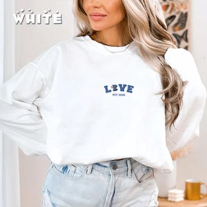 May include: White sweatshirt with the word "WHITE" printed in a stylized font at the top left. The word "LOVE" is printed in blue with a floral design and the text "EST 2026" below it. The sweatshirt is being worn by a person.