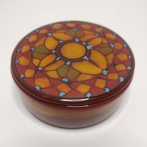 Könnte beinhalten: Eine runde Keramik-Schmuckdose mit Deckel. Der Deckel hat ein geometrisches Design in Orange-, Rot- und Gelbtönen mit blauen Akzenten.