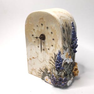 Può includere: Un orologio in ceramica bianca con un design floreale. L'orologio ha un quadrante bianco con lancette nere e un design floreale attorno al bordo. I fiori sono in tonalità di viola, blu e bianco.