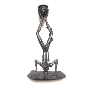 May include: Bronze sculpture of a figure in a handstand, holding a pot overhead. The figure has a detailed face and is atop a textured base. The sculpture is a dark bronze color.