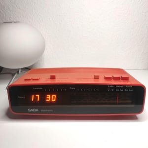 May include: A vintage SABA electronic alarm clock in a vibrant orange hue. The digital display shows the time as 17:30. Features include volume and radio controls. A retro design for a classic aesthetic.