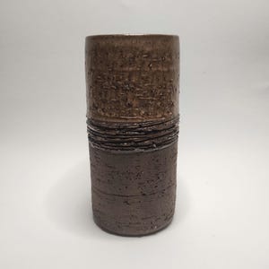 Könnte beinhalten: Eine braune Keramikvase mit einer strukturierten Oberfläche und einem Band aus dunkelbrauner Glasur um die Mitte.