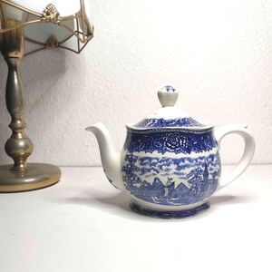 May include: White ceramic teapot with a blue and white landscape pattern. The teapot features a curved spout, a handle, and a lid with a small knob. The design includes floral and scenic elements.
