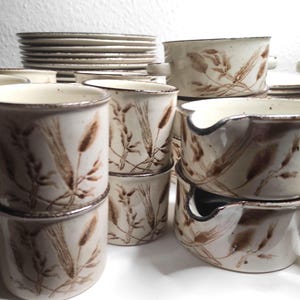 Op de afbeelding: Een verzameling beige keramisch servies met een tarwe-aren-ontwerp in bruin. De set bevat mokken, kommen en borden. De randen van de stukken hebben een donkerbruine rand.