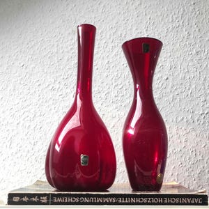 Può includere: Due vasi di vetro rosso rubino di forme diverse. Il vaso a sinistra ha una base bulbosa e un collo lungo. Il vaso a destra ha una parte superiore svasata. Entrambi i vasi hanno una finitura lucida e sono appoggiati su un libro.