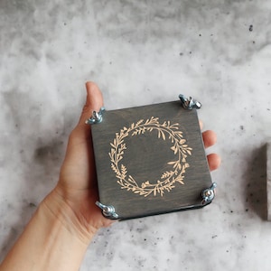 May include: A square, dark gray wooden box with a floral wreath design etched on the top. The box is held by a hand and secured with four metal wing nuts. A small concrete planter with greenery is to the right.