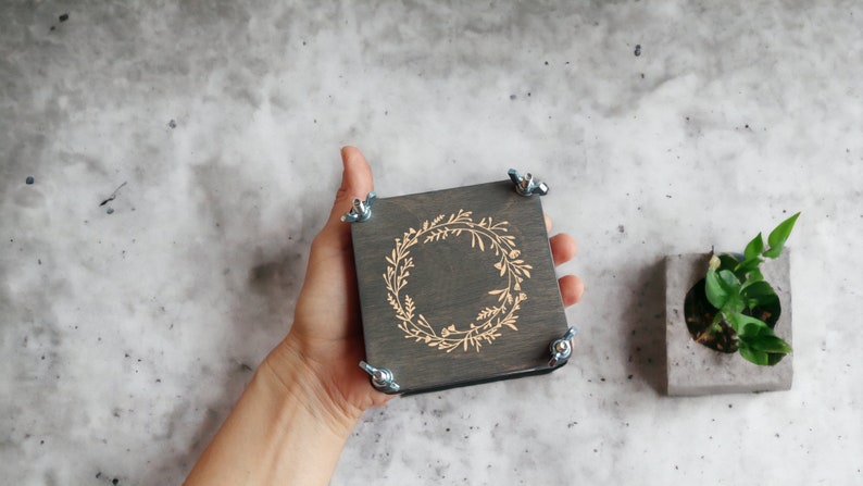 May include: A square, dark gray wooden box with a floral wreath design etched on the top. The box is held by a hand and secured with four metal wing nuts. A small concrete planter with greenery is to the right.