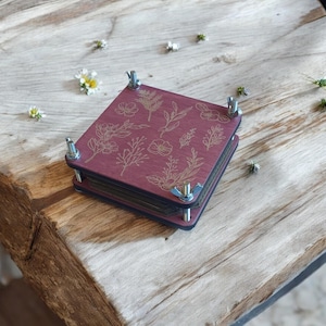 Könnte beinhalten: Eine quadratische Blumenpresse aus dunkelmagentafarbenem Holz mit eingravierten botanischen Illustrationen verschiedener Blumen und Blätter. Die Presse verfügt über vier Metallschrauben an den Ecken mit silbernen Flügelmuttern zum Festziehen.