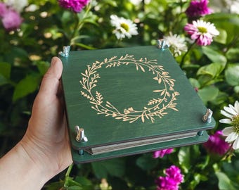 May include: A square, dark green wooden flower press featuring a laser-engraved floral wreath design on the top cover. The device is held together by four metal bolts with wing nuts at each corner to secure the contents inside.