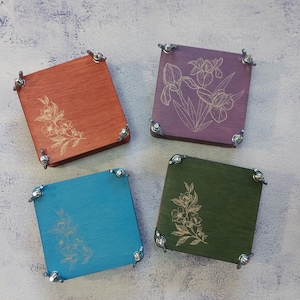 May include: Four square wooden coasters in orange, purple, teal, and green, each with a floral design. The coasters have metal corner accents. The floral designs are etched into the wood.