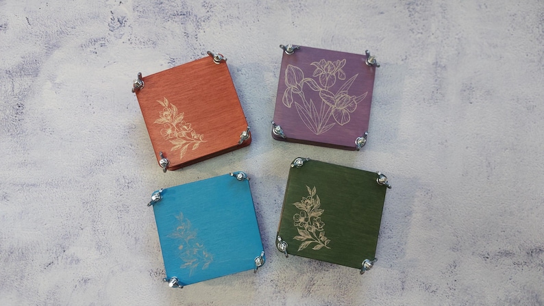 May include: Four square wooden coasters in orange, purple, teal, and green, each with a floral design. The coasters have metal corner accents. The floral designs are etched into the wood.