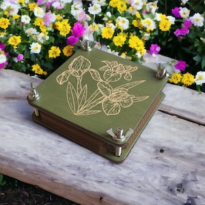 May include: A green wooden flower press with a floral design on the top. The press is secured with four wing nuts. The background features a wooden surface and colorful flowers.