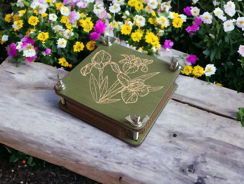 May include: A green wooden flower press with a floral design on the top. The press is secured with four wing nuts. The background features a wooden surface and colorful flowers.