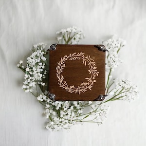 May include: A square, dark brown wooden box with a laser-etched floral wreath design on the lid. The box has metal corner protectors. The box is surrounded by white baby's breath flowers.