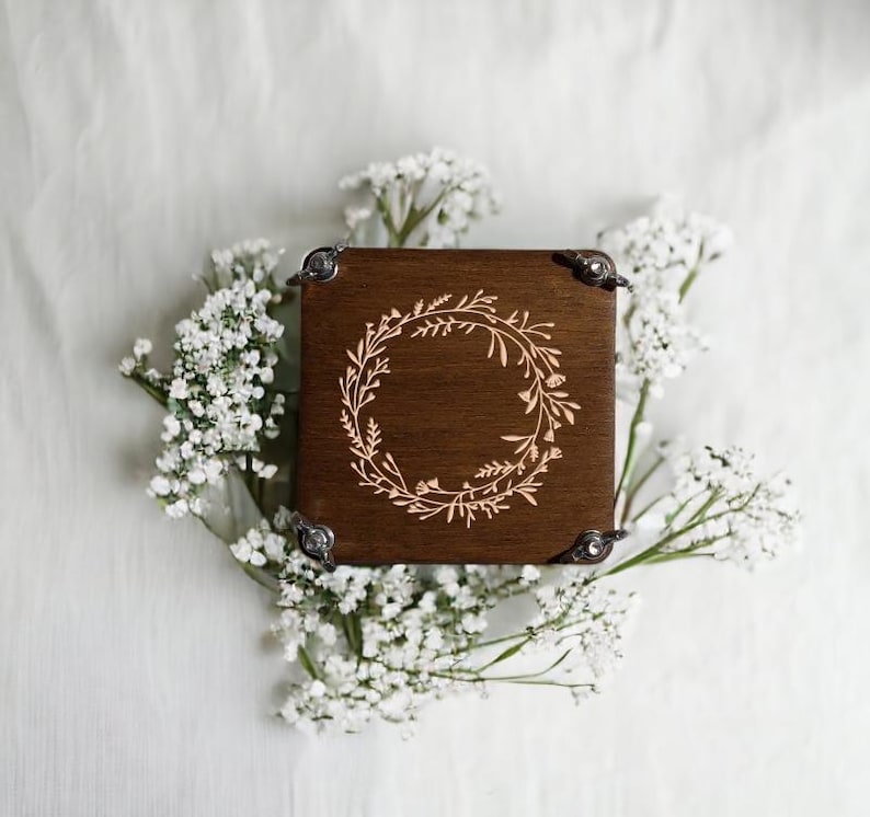 May include: A square, dark brown wooden box with a laser-etched floral wreath design on the lid. The box has metal corner protectors. The box is surrounded by white baby's breath flowers.