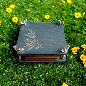 May include: A square, blue wooden flower press with silver wing nuts at each corner. The top of the press features a floral design. The press is sitting on a bed of green grass with yellow flowers.